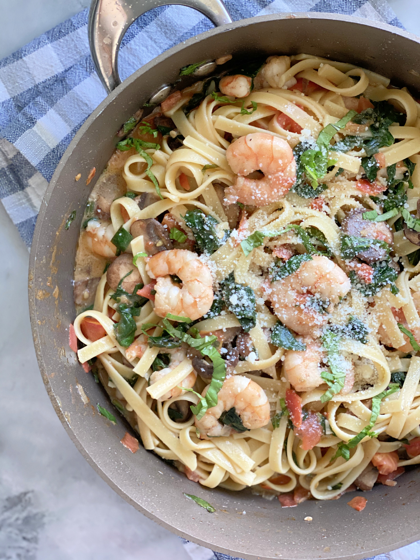 Brown pot filled with pasta, mushrooms, spinach, and shrimp on a blue and white checkered cloth.